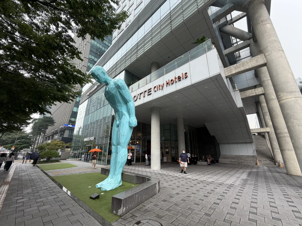 韓国・ソウルのロッテシティホテル明洞の外観とエントランス。ガラス張りの建物前に大きな青緑色の人物オブジェが立つ様子